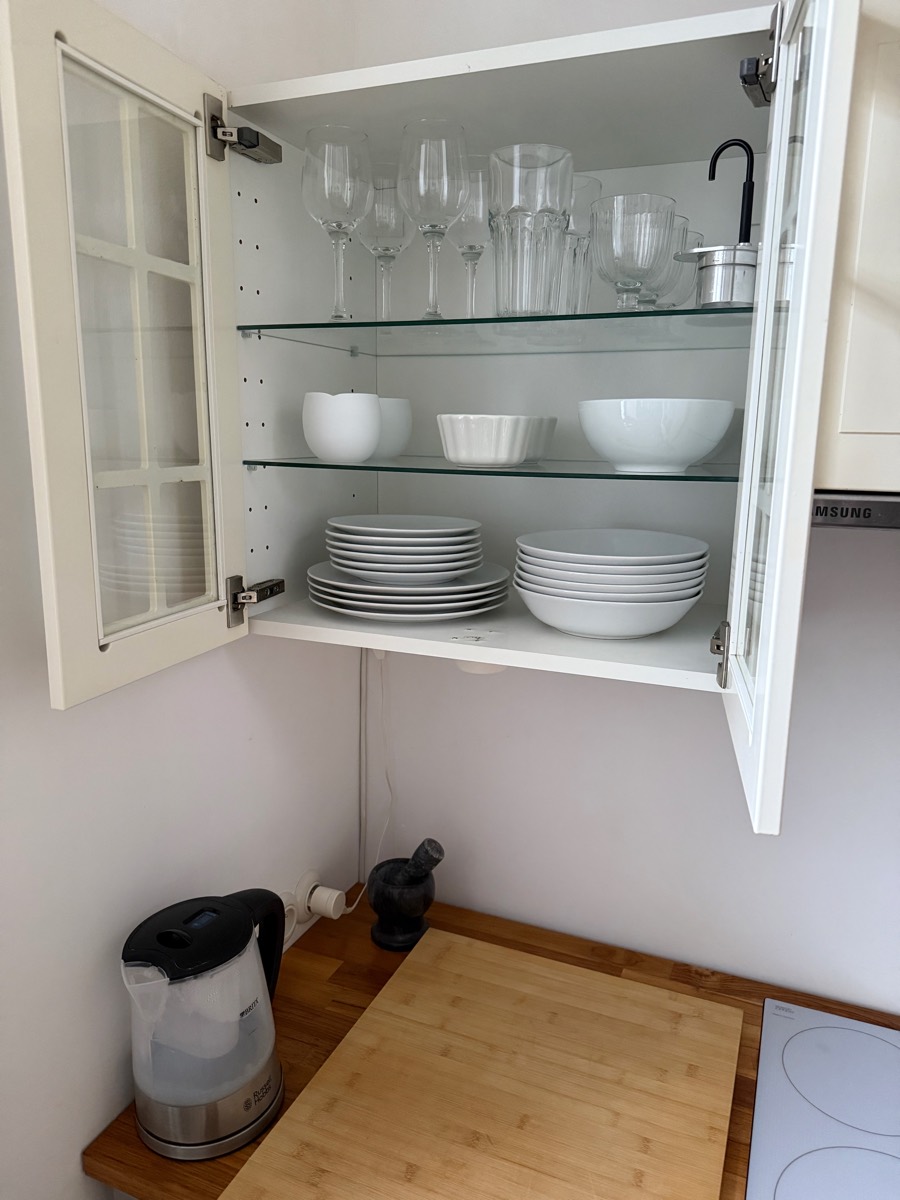 Open glass-front cabinet with wine glasses, tumblers, bowls and plates above the kitchen worktop