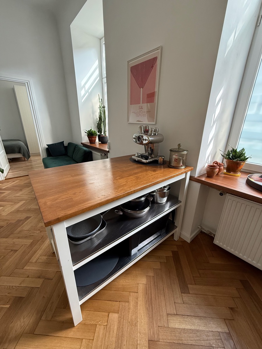 Oak-topped kitchen island with pans on the open shelf, looking through to the green sofa and the bedroom